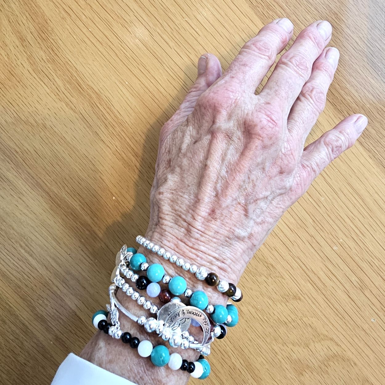 Hand wearing a collection of Bessie Bae faith and spirituality bracelets with silver charms and turquoise gemstones on a wooden surface.