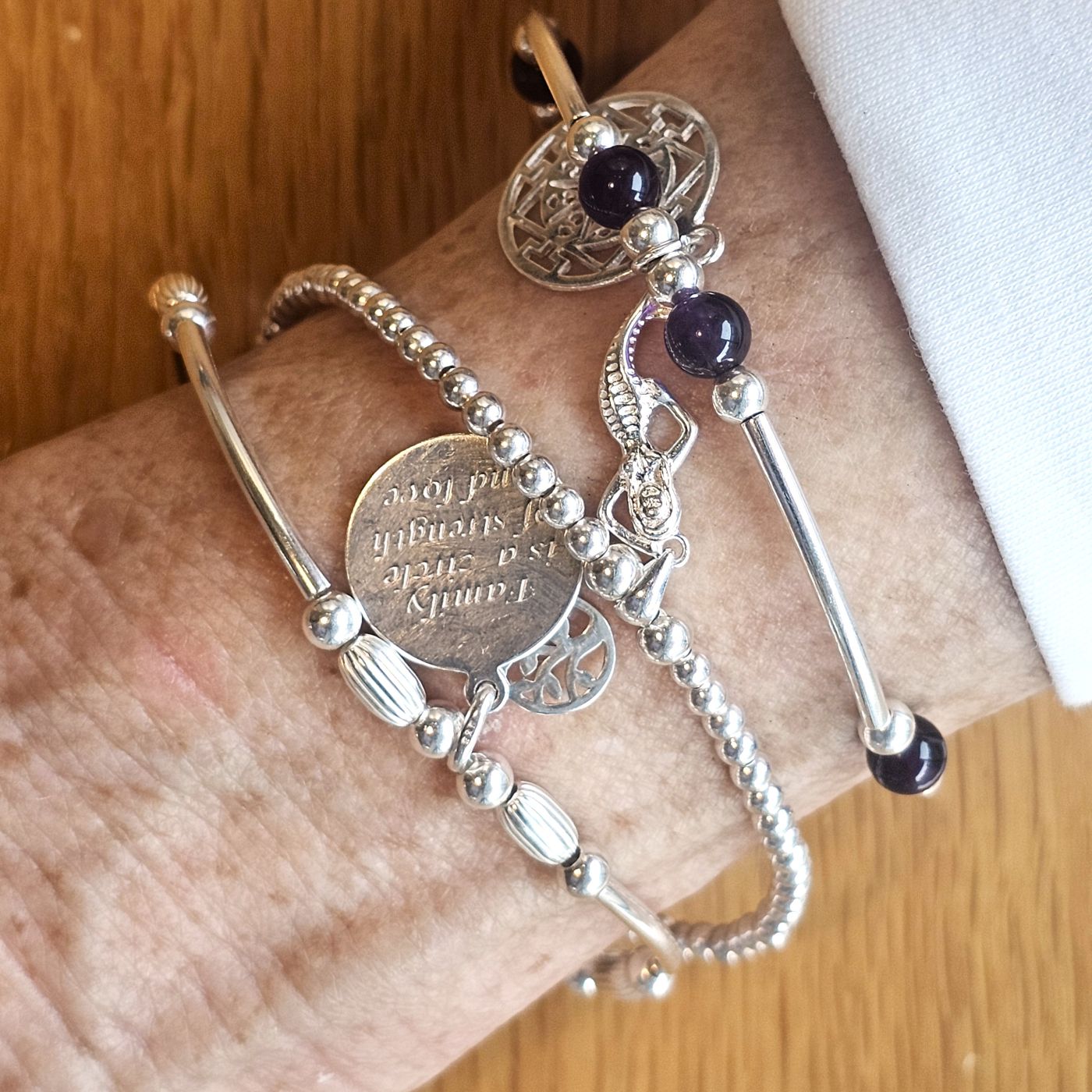 Wrist wearing Bessie Bae sterling silver charm bracelets with beaded and engraved details on a wooden surface.