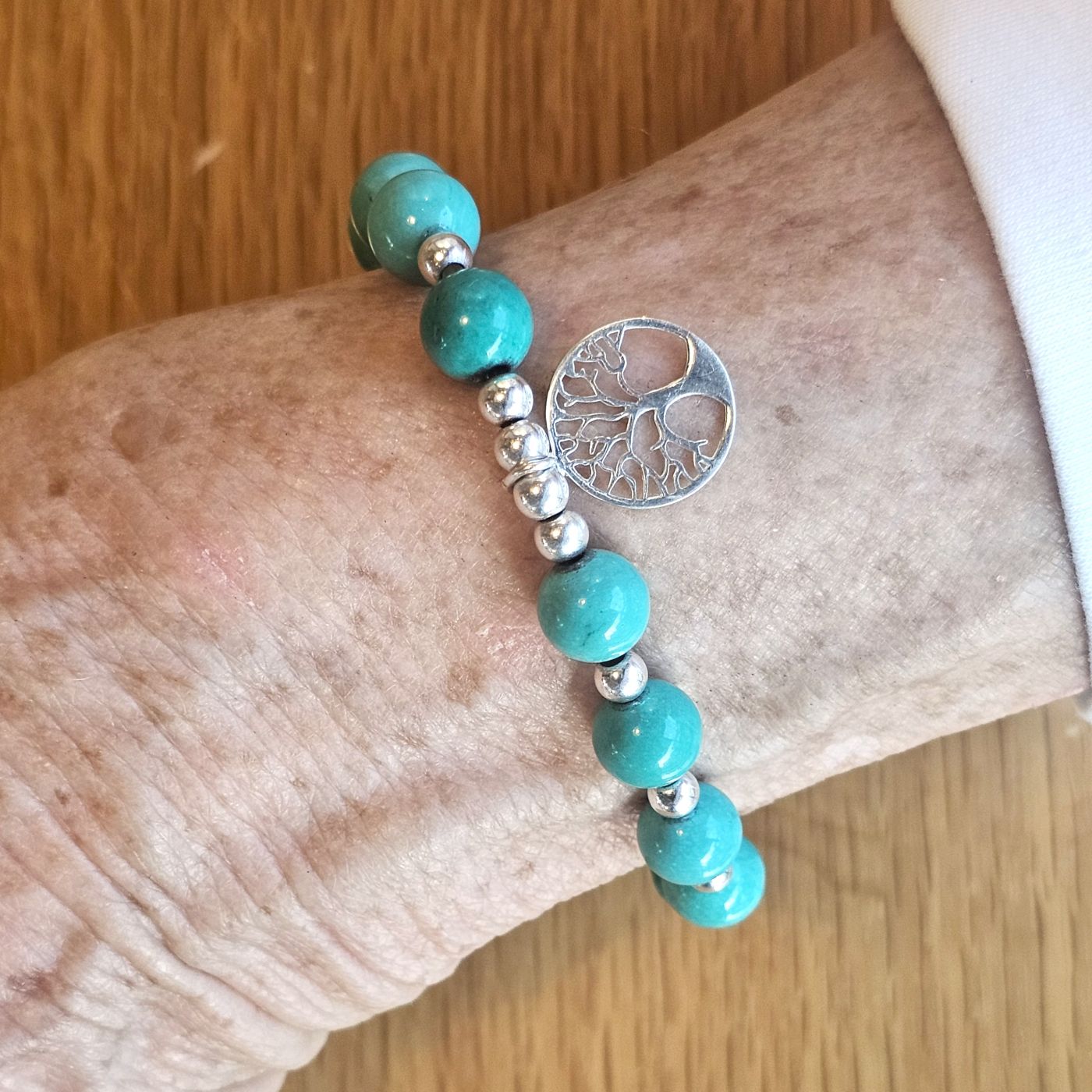 Wrist wearing a Bessie Bae turquoise beaded bracelet with a silver tree of life charm on a wooden surface.