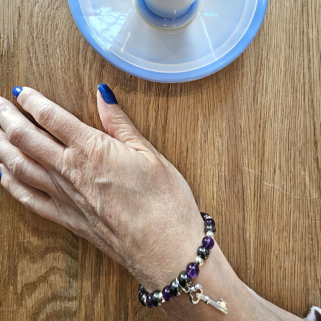 Amethyst and Hematite bracelet with Sterling Silver key charm from Bessie Bae worn on wrist while holding a blue cup on wooden table.