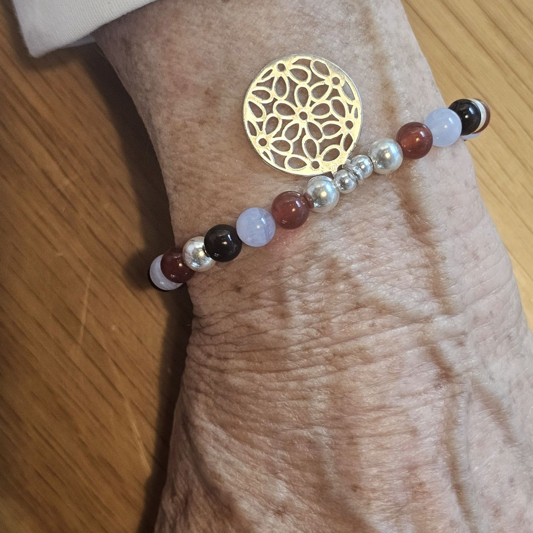 Wrist wearing a Bessie Bae bracelet with a gold floral disc charm and mixed gemstone beads in red, blue, black, and silver tones on a wooden surface.