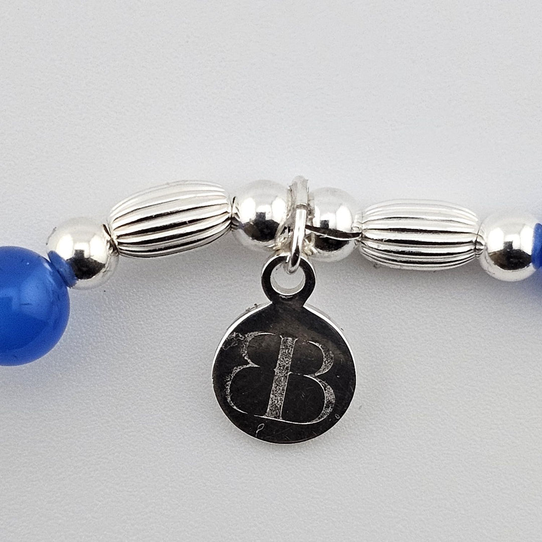Bessie Bae logo tag attached to silver sailboat charm bracelet with blue beads.
