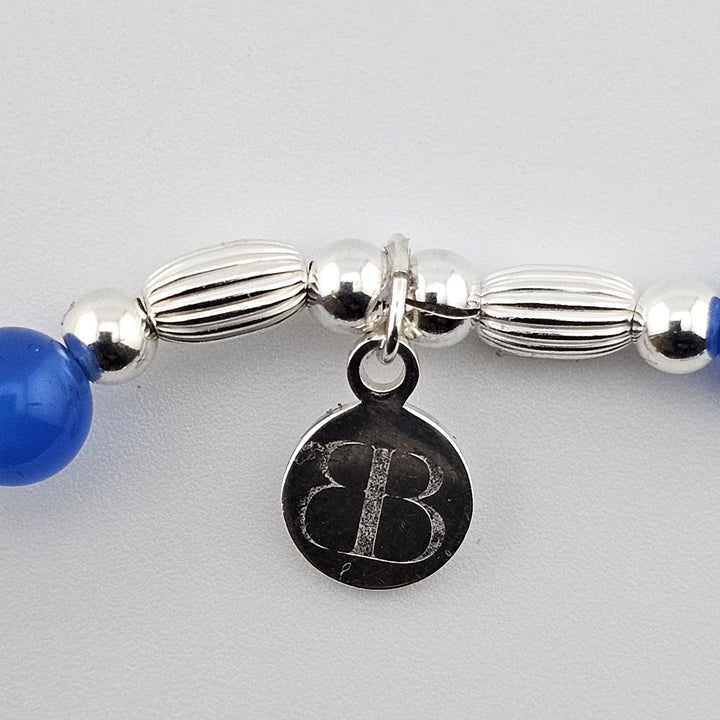 Bessie Bae logo tag attached to silver sailboat charm bracelet with blue beads.