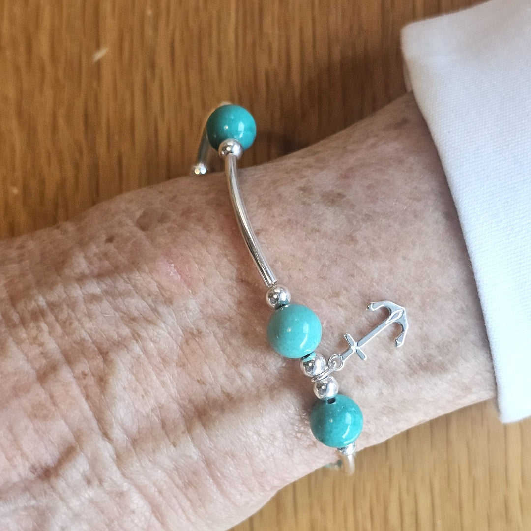 Wrist wearing a Bessie Bae silver anchor charm bracelet with turquoise beads on a wooden surface.