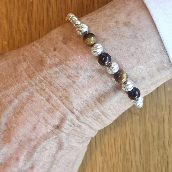 Wrist wearing a Bessie Bae silver and tiger’s eye beaded bracelet on a wooden surface.