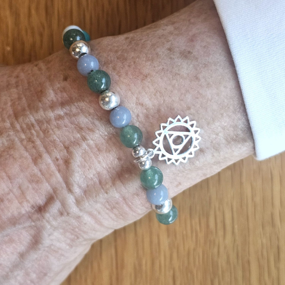 Wrist wearing a Bessie Bae silver chakra symbol bracelet with green and blue gemstone beads on a wooden surface.