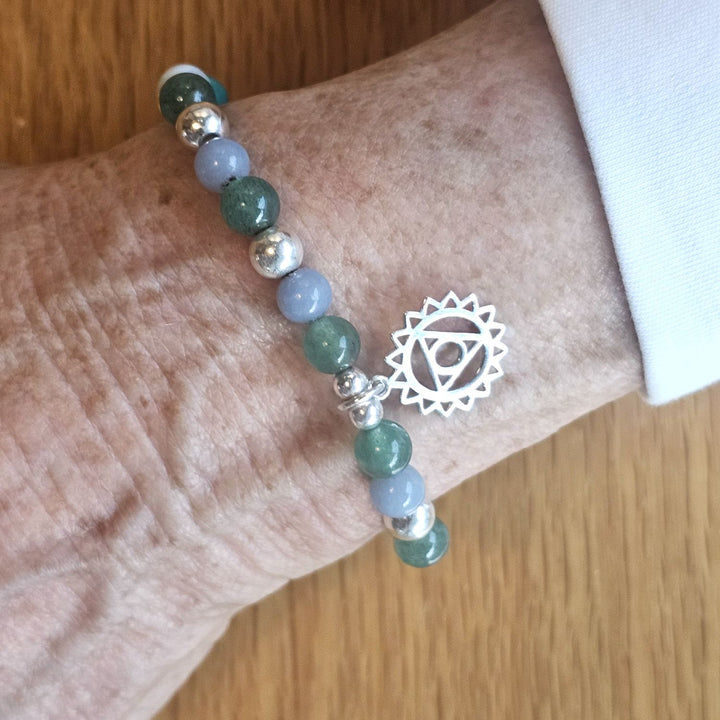 Wrist wearing a Bessie Bae silver chakra symbol bracelet with green and blue gemstone beads on a wooden surface.