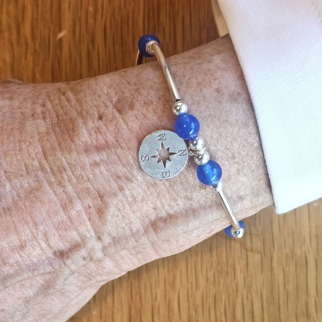 Wrist wearing a Bessie Bae silver compass charm bracelet with blue beads on a wooden surface.