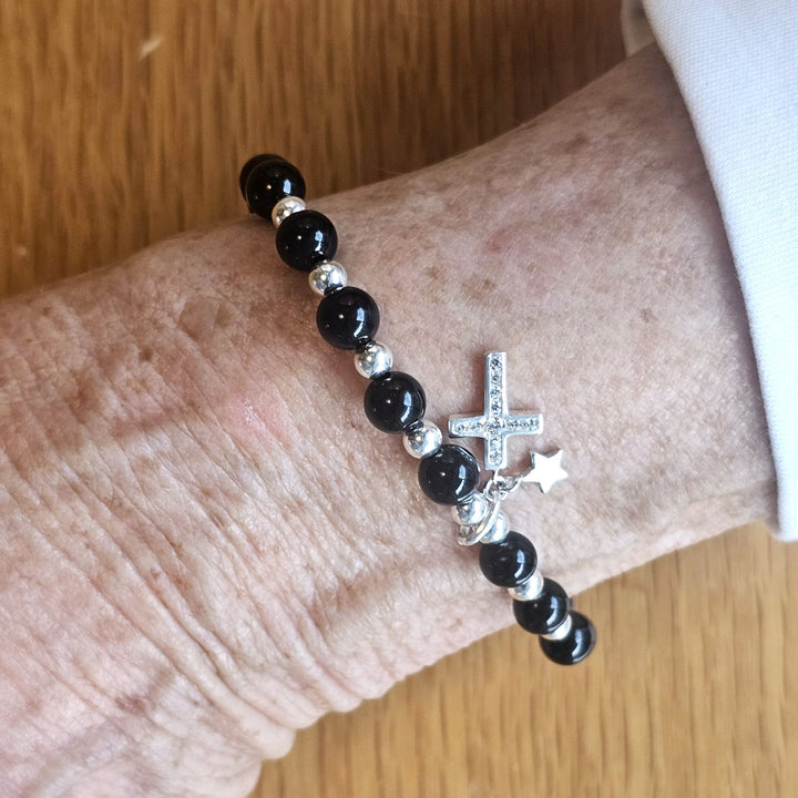Wrist wearing a Bessie Bae silver cross and star charm bracelet with black beads on a wooden surface.