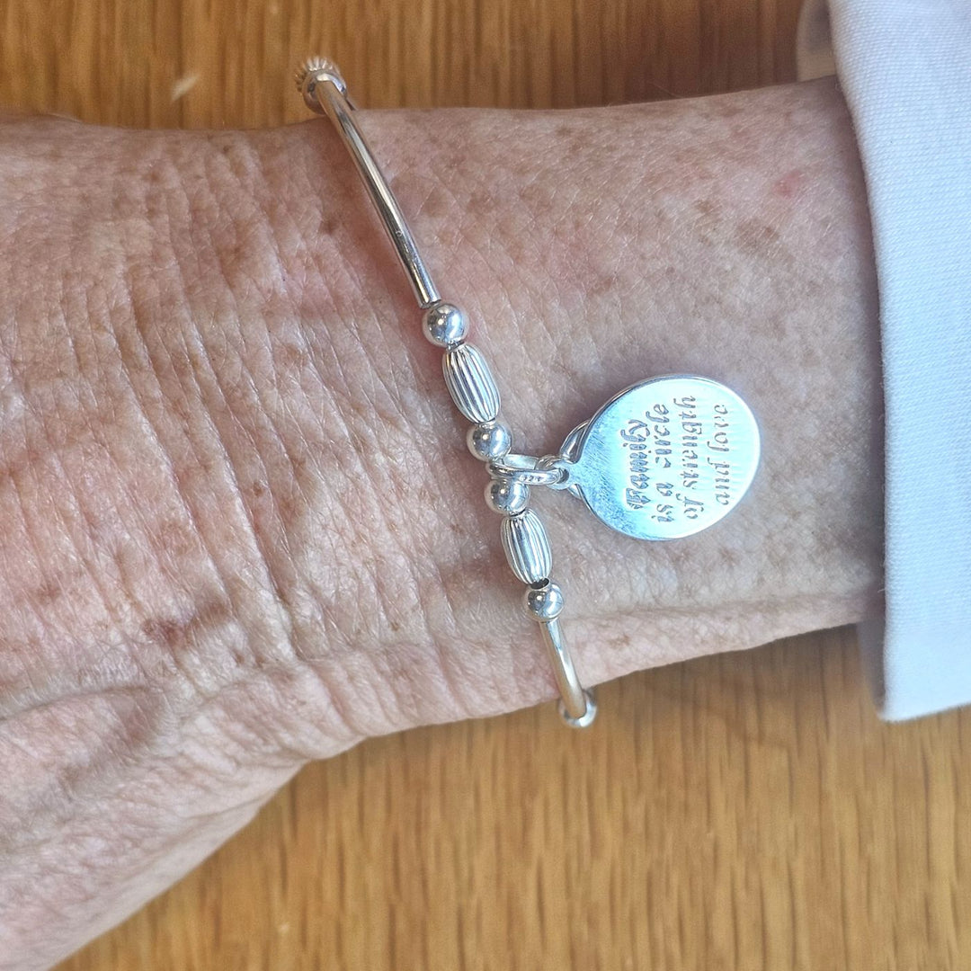 Wrist wearing a Bessie Bae silver family tree engraved charm bracelet with smooth and ribbed silver beads on a wooden surface.