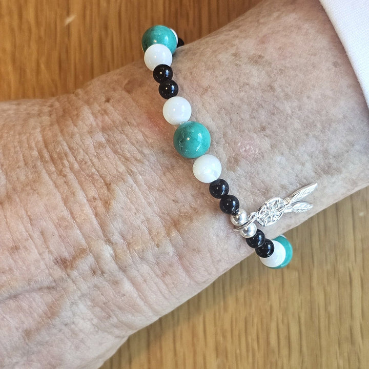 Wrist wearing a Bessie Bae silver feather charm bracelet with turquoise, white, and black beads on a wooden surface.