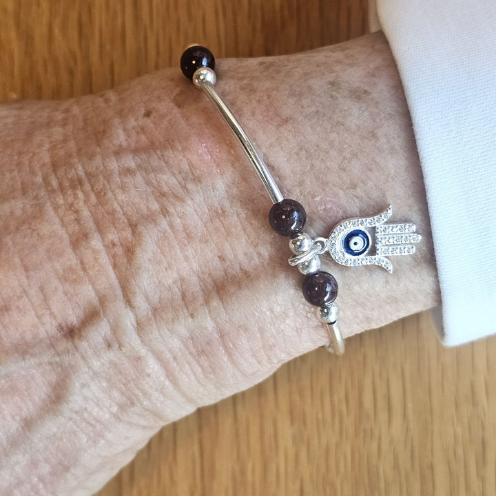 Wrist wearing a Bessie Bae silver Hamsa Hand charm bracelet with garnet beads on a wooden surface.