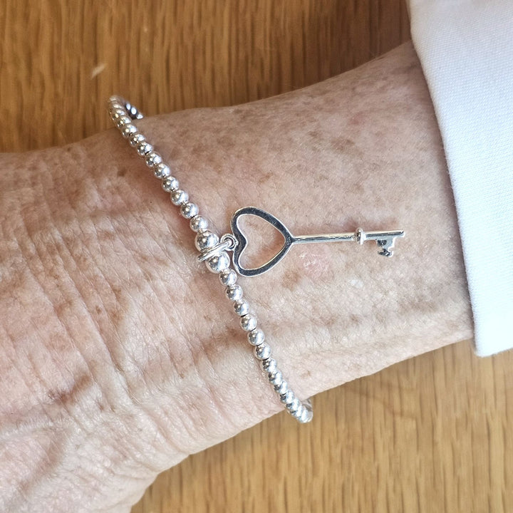 Wrist wearing a Bessie Bae silver heart key charm beaded bracelet on a wooden surface.
