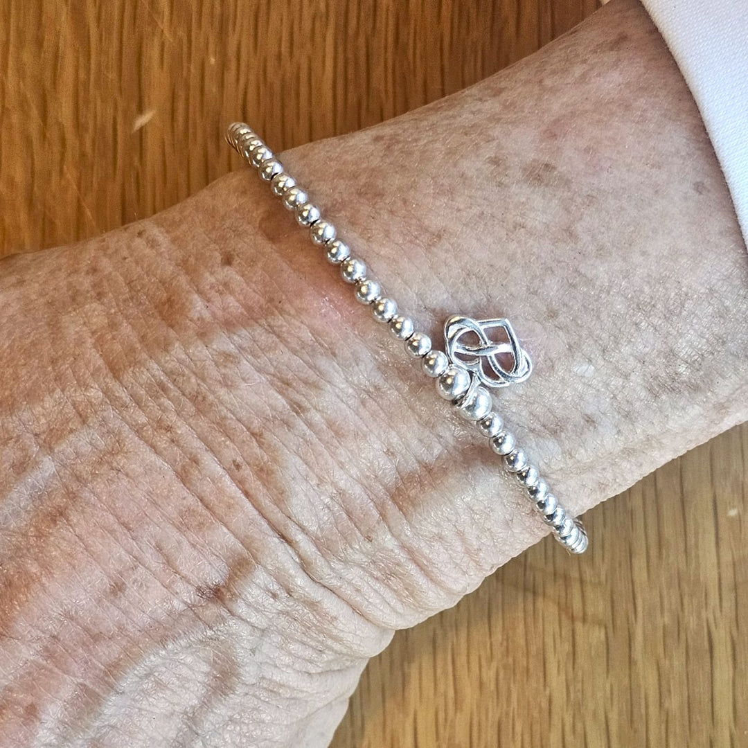 Wrist wearing a Bessie Bae silver infinity charm beaded bracelet on a wooden surface.