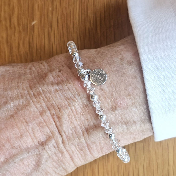 Wrist wearing a Bessie Bae silver logo charm bracelet with clear crystal beads on a wooden surface.