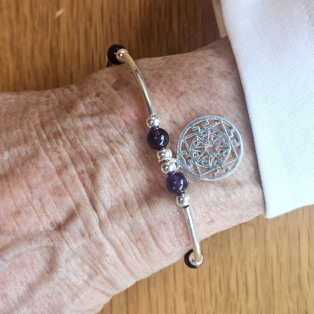 Wrist wearing a Bessie Bae silver mandala charm bracelet with amethyst and silver beads on a wooden surface.