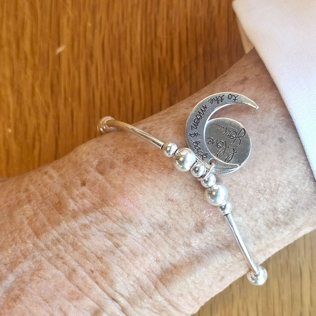 Wrist wearing a Bessie Bae silver bracelet with moon and back charm on a wooden surface.