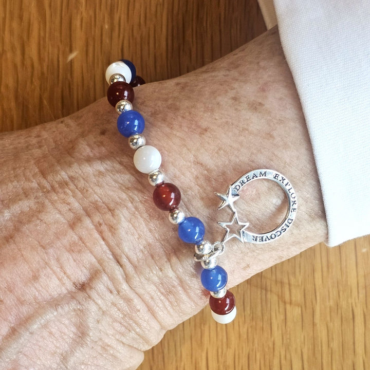 Wrist wearing a Bessie Bae silver moon and stars charm bracelet with red, blue, and white beads on a wooden surface.