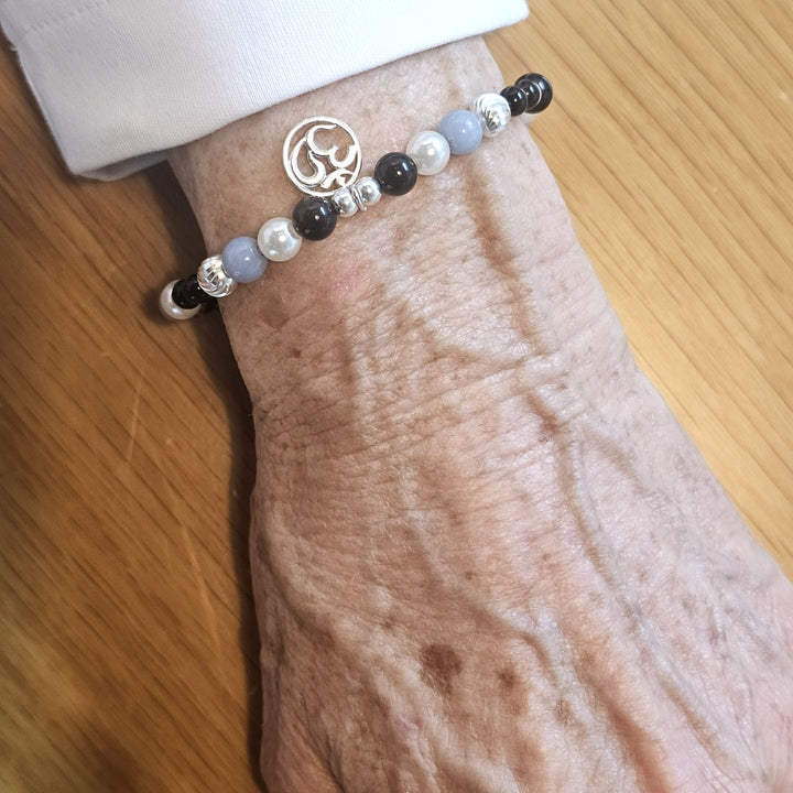 Wrist wearing a Bessie Bae silver Om charm bracelet with black, pearl, and blue beads on a wooden surface.