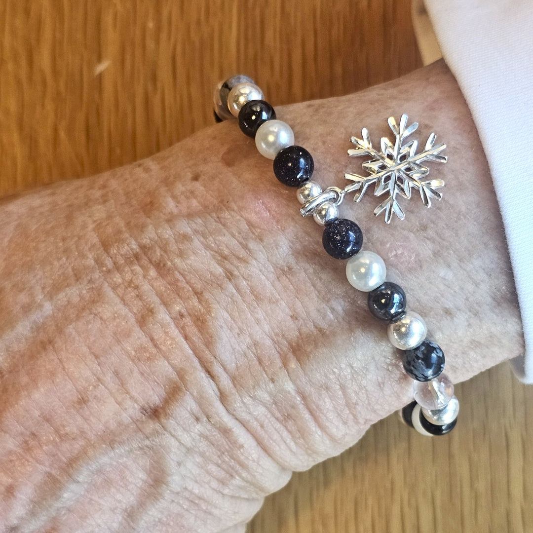 Wrist wearing a Bessie Bae silver snowflake charm bracelet with black, pearl, and silver beads on a wooden surface.