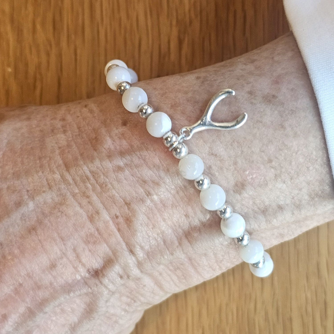 Wrist wearing a Bessie Bae silver wishbone charm bracelet with white beads on a wooden surface.
