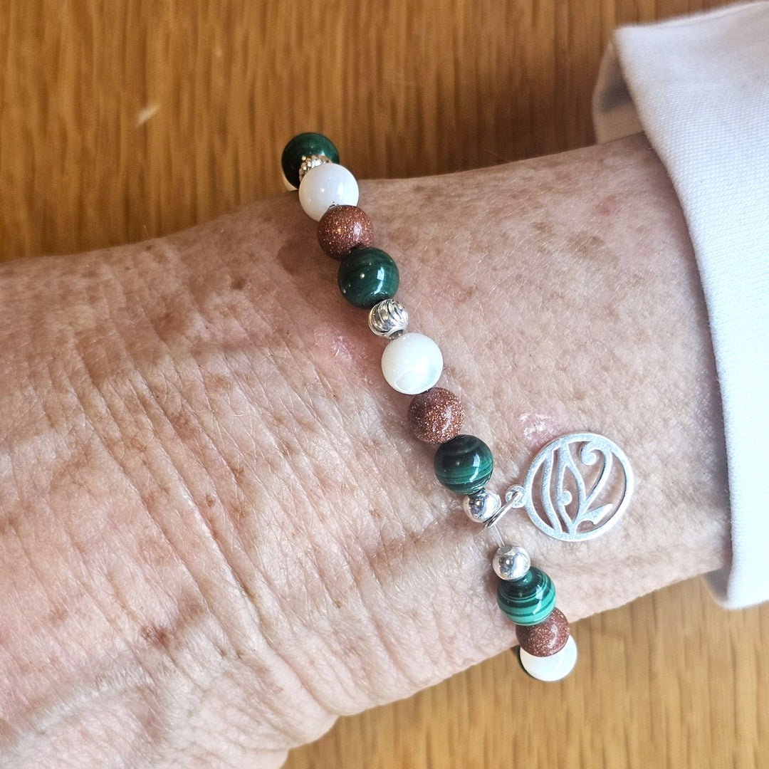 Wrist wearing a Bessie Bae silver zodiac charm bracelet with green, goldstone, and white beads on a wooden surface