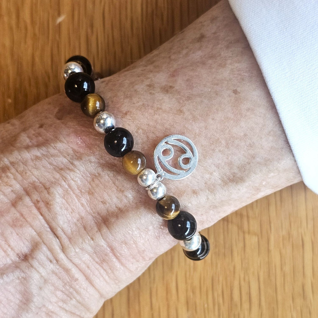 Wrist wearing a Bessie Bae silver zodiac charm bracelet with tiger’s eye, black, and silver beads on a wooden surface.