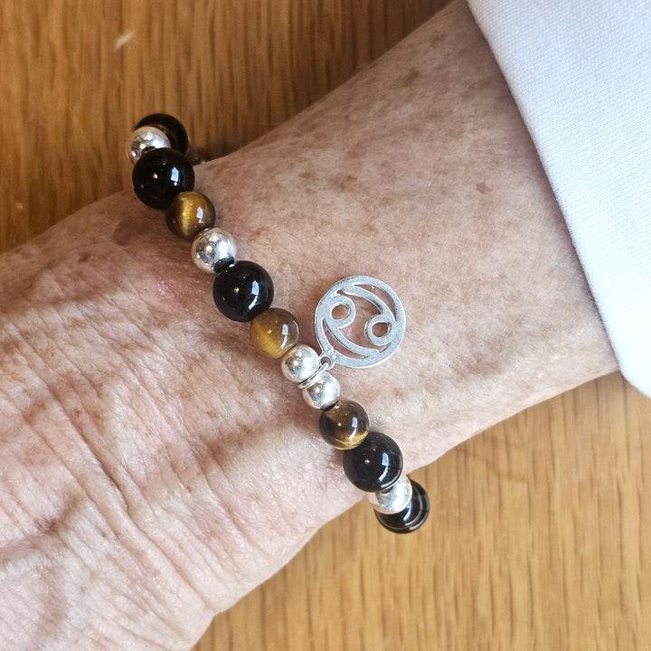 Wrist wearing a Bessie Bae silver zodiac charm bracelet with tiger’s eye, black, and silver beads on a wooden surface.