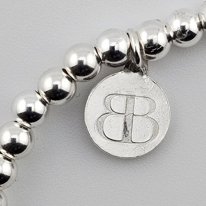 Bessie Bae silver logo charm attached to sterling silver beaded bracelet.