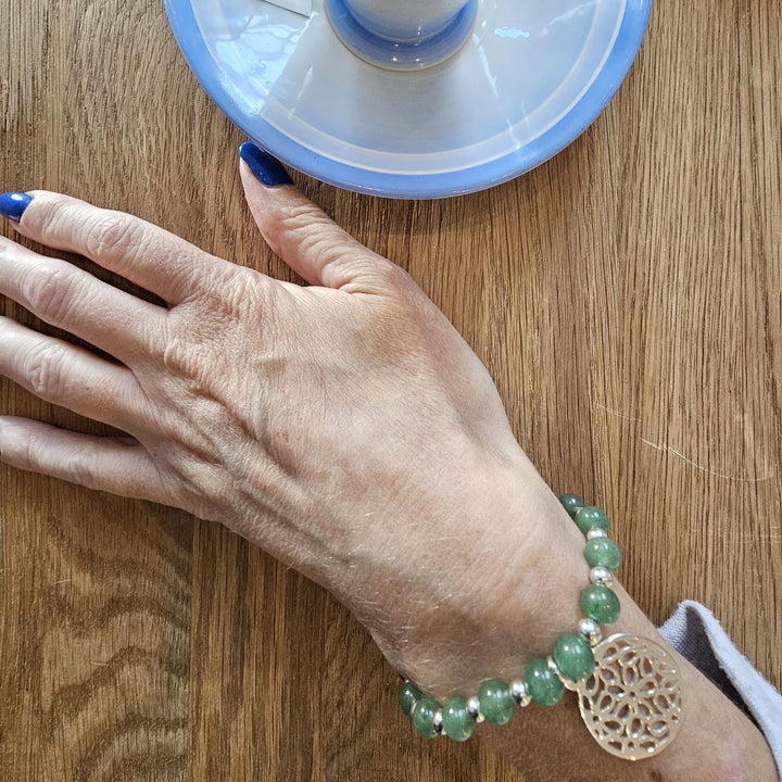 Green Aventurine bracelet with Sterling Silver filigree charm from Bessie Bae worn on wrist while holding a blue cup on wooden table.