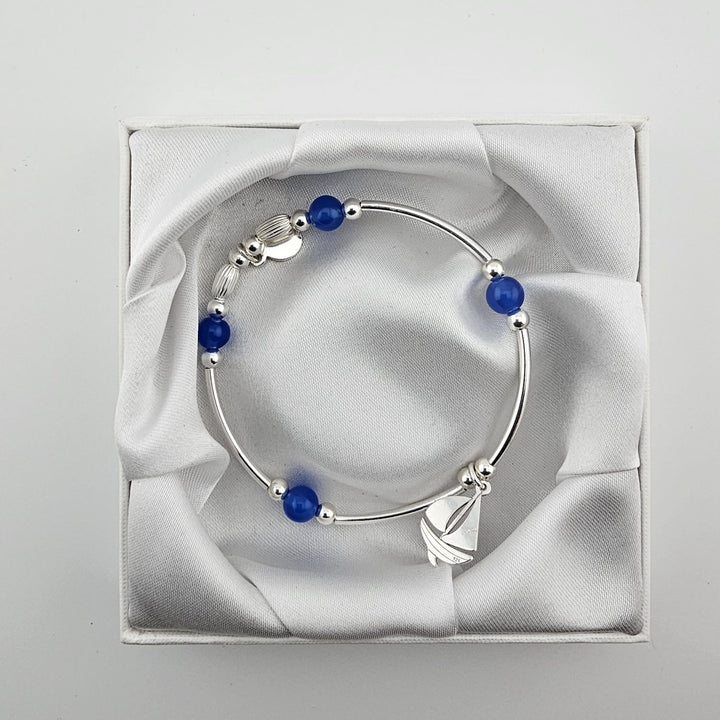 Top view of silver bracelet with blue beads and sailboat charm in white box.