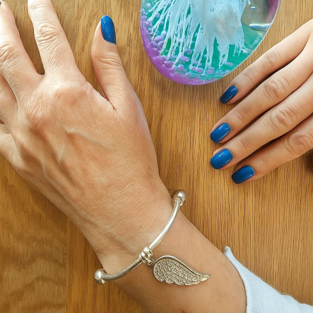 Sterling silver bracelet with angel wing charm, shown on wrist beside a lilac and green glass paperweight.