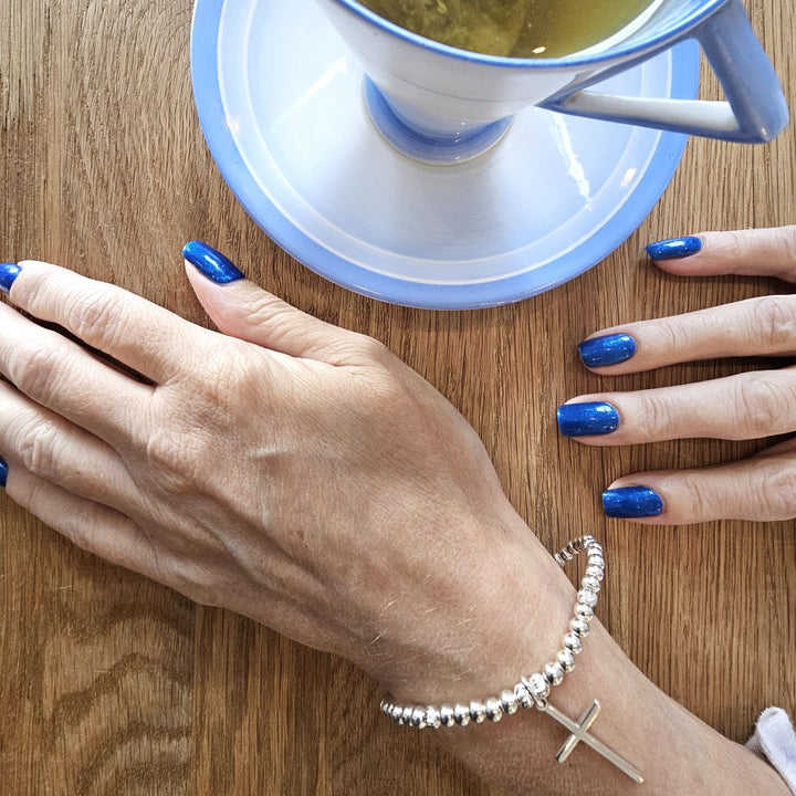 Sterling Silver Cross bracelet from Bessie Bae worn on wrist while holding a blue cup on a wooden table.