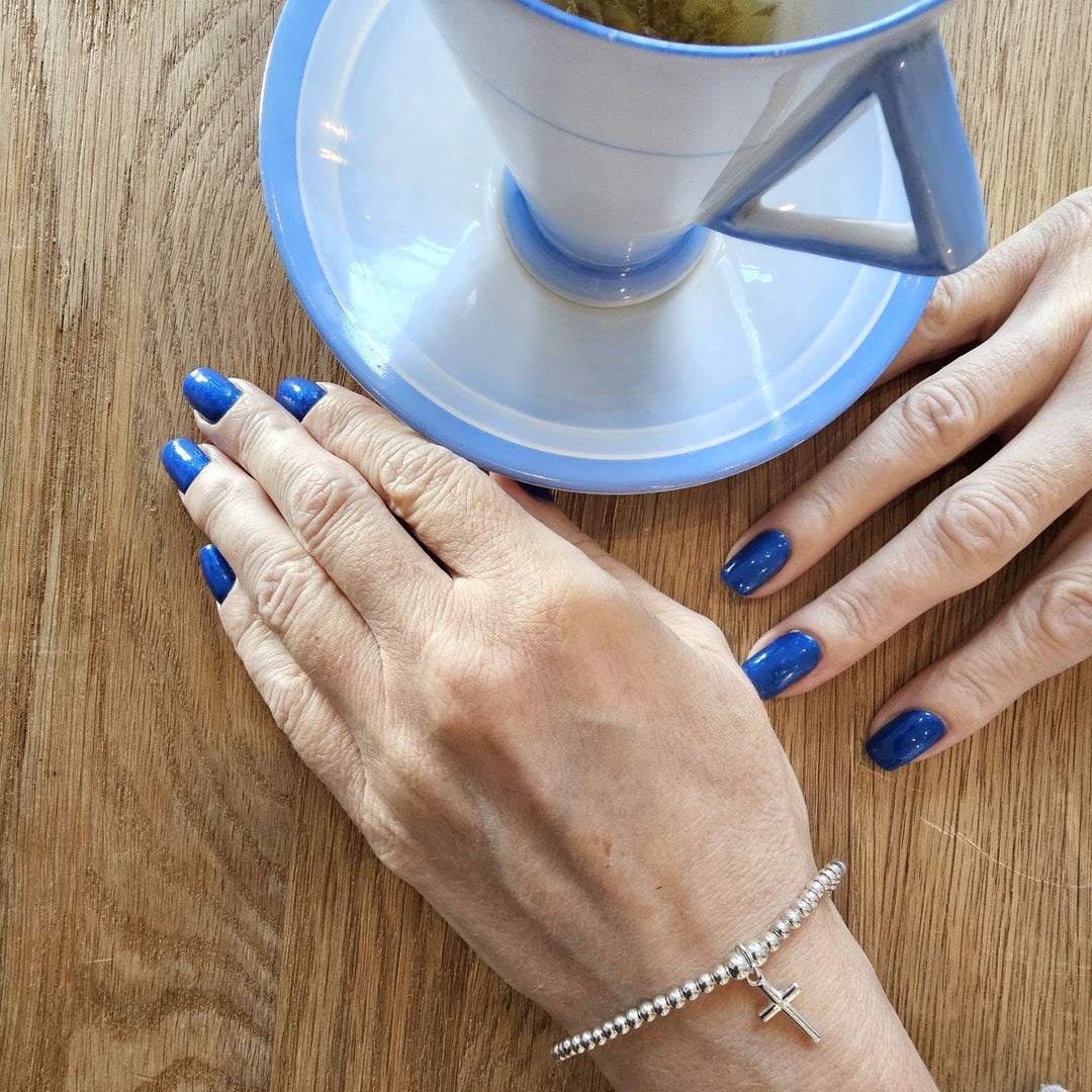 A person wearing a delicate sterling silver beaded bracelet with a cross charm, holding a cup of herbal tea on a wooden table.