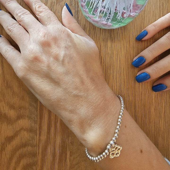 A person wearing a delicate sterling silver beaded bracelet with interlocking hearts charm, resting their hand beside a glass paperweight on a wooden table.