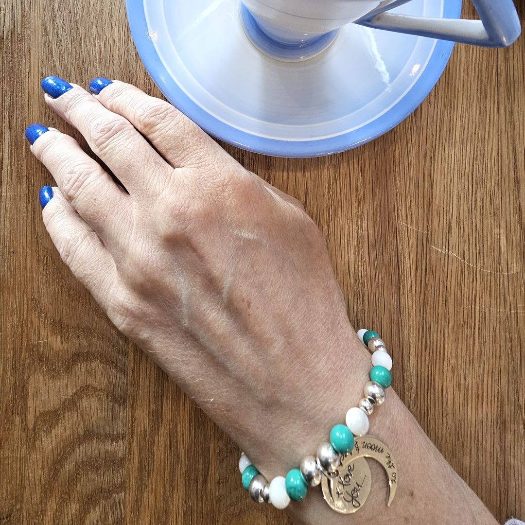 A person wearing a Sterling Silver bracelet with turquoise and mother of pearl beads, featuring an engraved crescent moon charm that reads “I love you to the moon and back,” while holding a cup of herbal tea.
