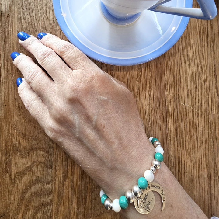 A person wearing a Sterling Silver bracelet with turquoise and mother of pearl beads, featuring an engraved crescent moon charm that reads “I love you to the moon and back,” while holding a cup of herbal tea.